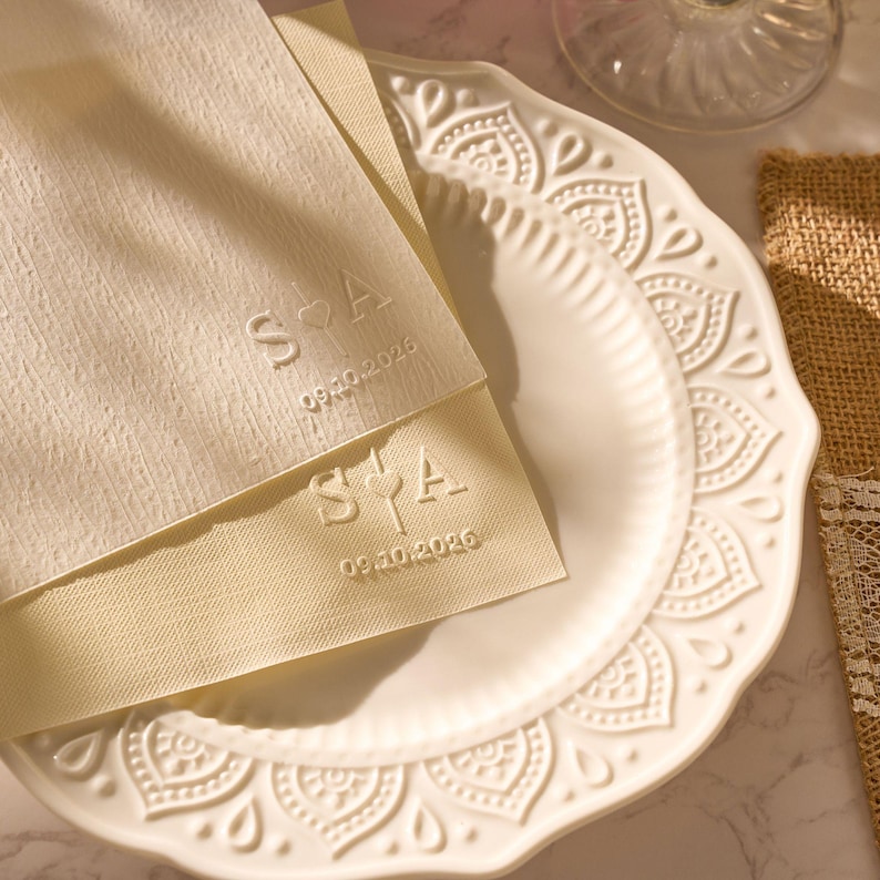 May include: Two cream-coloured napkins with embossed initials and a date, resting on a white decorative plate. The napkins feature the initials "S♡A" and the date "09.10.2026". The plate has a detailed, embossed pattern.