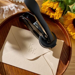 May include: A black letter seal tool is pressing down on a cream envelope, creating an embossed monogram. The envelope is on a wooden tray, with sunflowers in the background. The monogram appears to be the letters K and L.