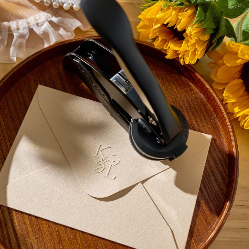 May include: A black letter seal tool is pressing down on a cream envelope, creating an embossed monogram. The envelope is on a wooden tray, with sunflowers in the background. The monogram appears to be the letters K and L.