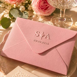 May include: A pale pink envelope featuring embossed initials "S&A" and the date "09.10.2026". The envelope is closed. Pink roses and a glass are in the background. The envelope is on a white surface.