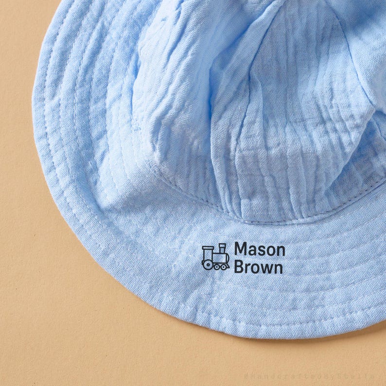 May include: A light blue baby sun hat with a soft, crinkled texture. The hat features the name "Mason Brown" printed in black, along with a small train graphic. The hat is designed to provide sun protection.