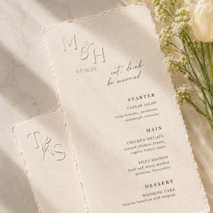 May include: Off-white wedding menu cards with rough edges. The cards feature embossed initials, the date 07.18.26, and the phrase "eat, drink, be married." The menu lists starter, main, and dessert courses.