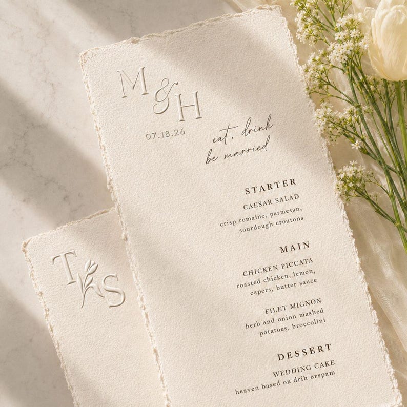 May include: Off-white wedding menu cards with rough edges. The cards feature embossed initials, the date 07.18.26, and the phrase "eat, drink, be married." The menu lists starter, main, and dessert courses.