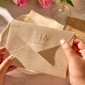 May include: A beige envelope with embossed initials "S&A" and the date "09.10.2026". The envelope is held by a person's hands, resting on a white plate with a floral design. A napkin and a piece of burlap are in the background.