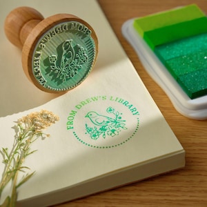 K&ouml;nnte beinhalten: Ein gr&uuml;ner und wei&szlig;er Stempel mit dem Text "From Drew's Library" und einer kleinen Vogel-Illustration. Der Stempel befindet sich auf einer wei&szlig;en Oberfl&auml;che mit einer kleinen gr&uuml;nen Pflanze.