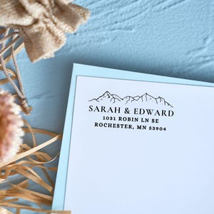 Pu&ograve; includere: Un set di cancelleria azzurro chiaro con una carta bianca con un disegno di catena montuosa e i nomi "SARAH & EDWARD" stampati in nero. Anche l'indirizzo 1031 ROBIN LN SE, ROCHESTER, MN 53904 &egrave; stampato.