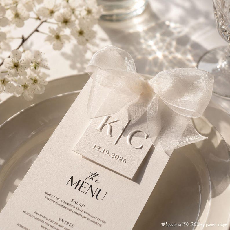 May include: A cream-coloured wedding menu card with a sheer white ribbon bow. The menu features the initials "K/C" and the date "12.19.2026". The menu is on a white plate, with white flowers in the background.