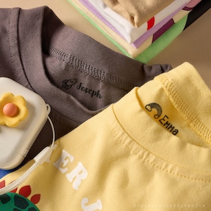 May include: Close-up of children's t-shirts in shades of gray and yellow, with the names "Joseph" and "Emma" printed on the collars. A stack of folded shirts in various colors is visible in the background. A white and yellow flower-shaped object is in the foreground.
