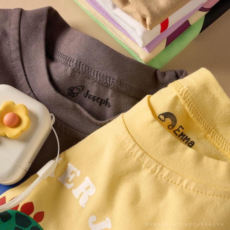 May include: Close-up of children's t-shirts in shades of gray and yellow, with the names "Joseph" and "Emma" printed on the collars. A stack of folded shirts in various colors is visible in the background. A white and yellow flower-shaped object is in the foreground.
