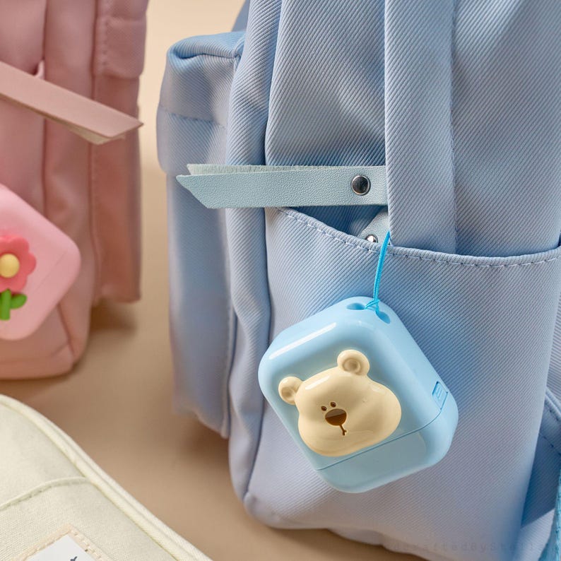 May include: A light blue backpack with a small, square, blue bear-shaped accessory hanging from it. The bear has a light brown face and a small, brown nose. A pink backpack and a cream-colored bag are also visible.