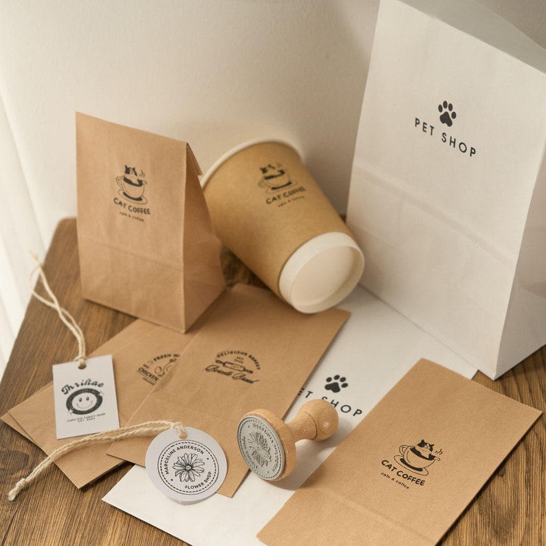 K&ouml;nnte beinhalten: Eine Sammlung brauner Papiert&uuml;ten mit verschiedenen Logos, darunter "Cat Coffee" und "Pet Shop". Es gibt auch zwei runde Gummistempel, einer mit einem Blumendesign und der andere mit einem Katzendesign. Die Stempel befinden sich auf einer wei&szlig;en Oberfl&auml;che.