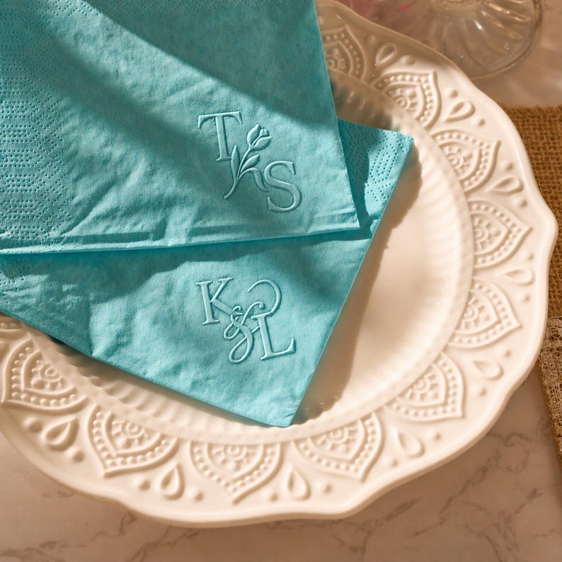 May include: Two folded, light blue napkins with embroidered initials sit on a white, decorative plate. The napkins feature the initials "TS" and "KL" in a delicate script. The plate has a scalloped edge and embossed floral patterns.