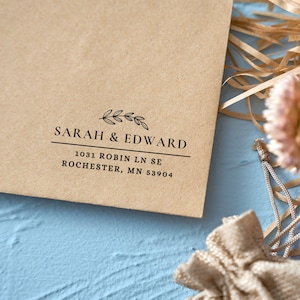 Pu&ograve; includere: Una busta di carta kraft con i nomi "SARAH & EDWARD" stampati in nero, insieme a un indirizzo. Un rametto decorativo di foglie &egrave; sopra i nomi. La busta &egrave; su una superficie blu con elementi decorativi.