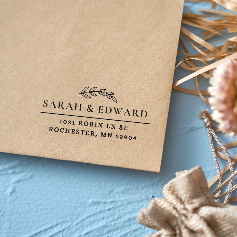 Pu&ograve; includere: Una busta di carta kraft con i nomi "SARAH & EDWARD" stampati in nero, insieme a un indirizzo. Un rametto decorativo di foglie &egrave; sopra i nomi. La busta &egrave; su una superficie blu con elementi decorativi.