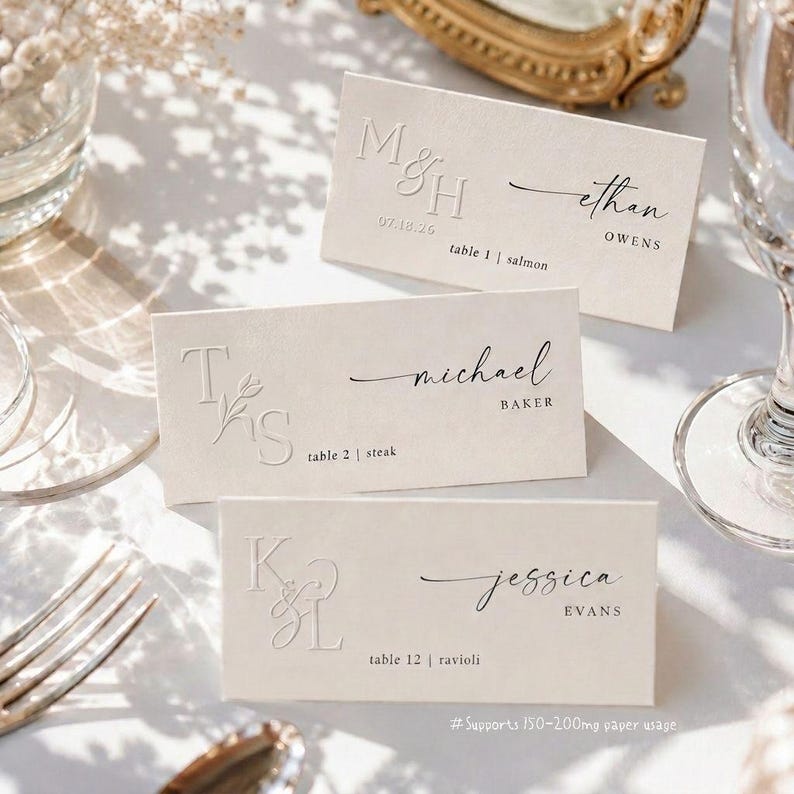 May include: Three cream-coloured place cards with embossed initials and names in elegant script. Each card includes table number and meal choice. The cards are arranged on a white surface, with a fork and glass visible.