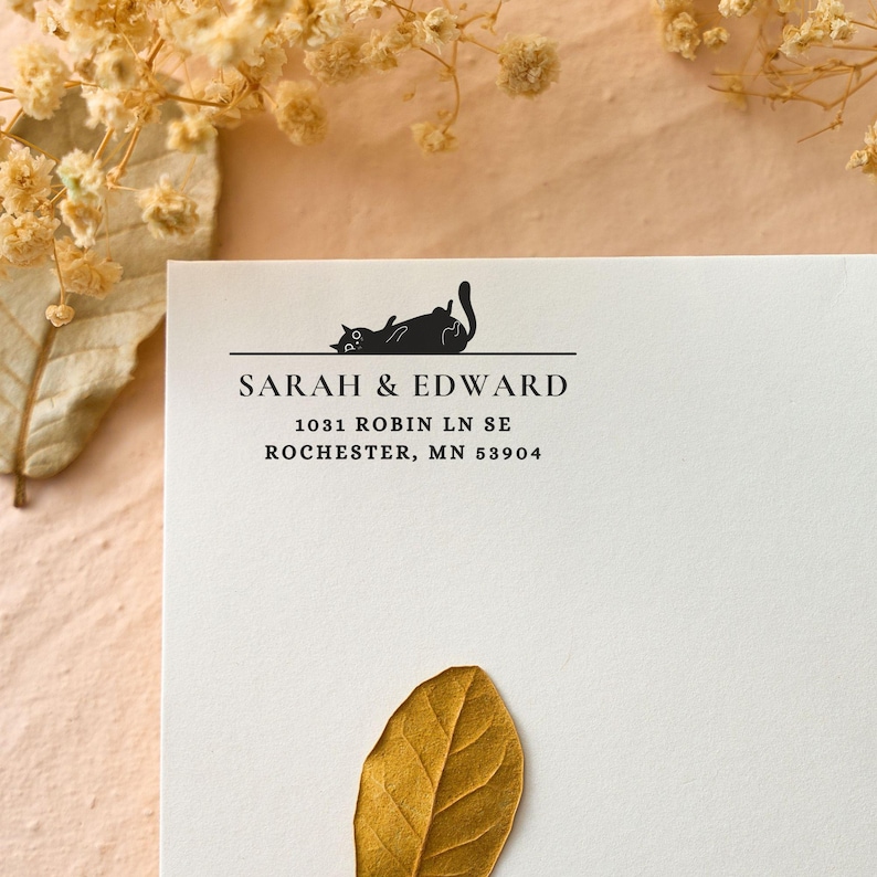 Pu&ograve; includere: Una carta bianca con testo nero e un'illustrazione di un gatto nero. Il testo recita "SARAH & EDWARD" sopra un indirizzo. Una foglia secca ed elementi floreali sono sullo sfondo.