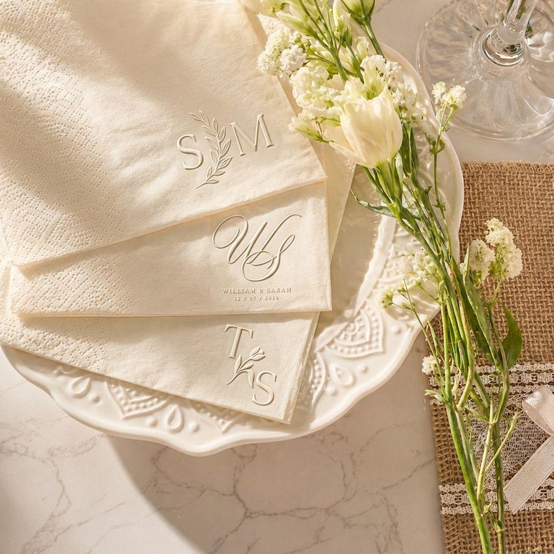 May include: Ivory-coloured personalised napkins with embossed initials and floral accents. The napkins are arranged on a white, decorative plate, alongside white flowers and a burlap placemat. The initials include "SM", "WS", and "TS".