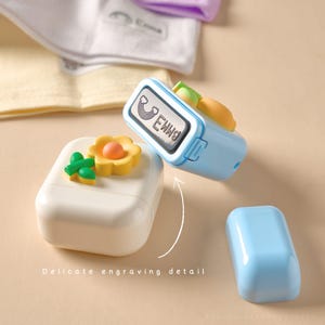 May include: A light blue stamp with a rainbow and the text "Ewng". A white rectangular case with a yellow flower and green leaves is next to the stamp. A light blue cap is also visible. The text "Delicate engraving detail" is at the bottom.