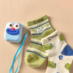 May include: A light blue, cartoon-faced label maker with a blue strap sits next to two pairs of children's socks. One pair is green and white striped with the name "Lucas" and the letter "M". The other pair is white with green dinosaur graphics and the name "Harper".