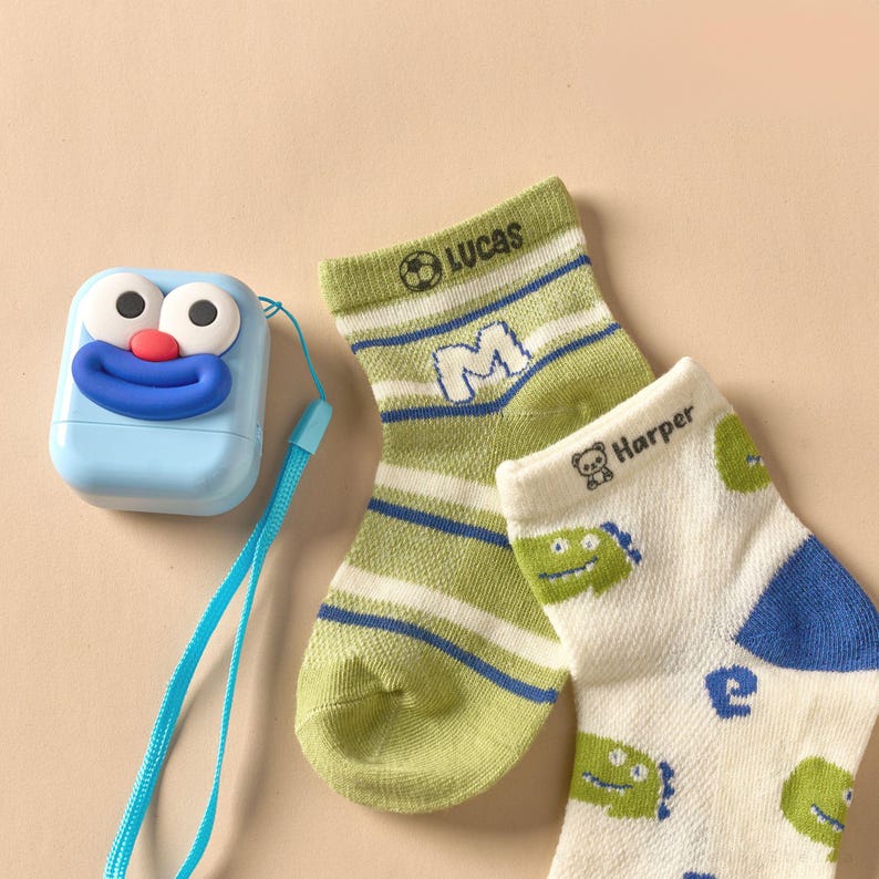 May include: A light blue, cartoon-faced label maker with a blue strap sits next to two pairs of children's socks. One pair is green and white striped with the name "Lucas" and the letter "M". The other pair is white with green dinosaur graphics and the name "Harper".