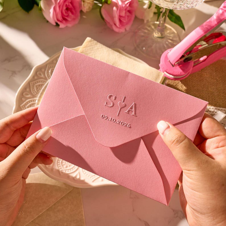 May include: A pink envelope with embossed initials and a date is held. The envelope is on a white plate with a floral design. The text on the envelope reads "S&A" and "09.10.2026".