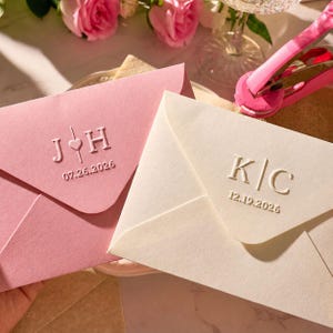 May include: Two envelopes, one light pink and one off-white, with embossed initials and dates. The pink envelope features "J&hearts;H" and "07.26.2026". The off-white envelope displays "K|C" and "12.19.2026". Suitable for wedding invitations or announcements.