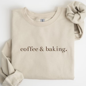 May include: A beige sweatshirt with the words "coffee & baking" printed in brown. The long sleeves are folded. The sweatshirt is made of a soft material and is laid flat on a white surface.