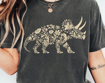 Puede incluir: Una camiseta de manga corta gris carbón con un gran gráfico de tricerátops relleno de un intrincado patrón floral y de enredaderas de color crema. La palabra PEPPER está impresa en la esquina superior derecha. La camiseta tiene cuello redondo.