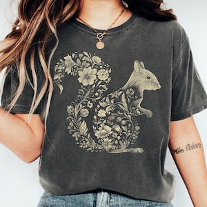 May include: Dark gray t-shirt featuring a detailed illustration of a squirrel composed of floral elements. The design is in a light beige color, creating a contrast against the shirt's darker hue. The shirt has a classic crew neck and short sleeves.