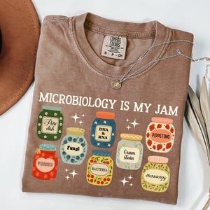 May include: A brown t-shirt with the text "MICROBIOLOGY IS MY JAM" and illustrations of jars labeled with microbiology terms like "Petri dish," "Fungi," and "Bacteria." A silver necklace is draped across the shirt.