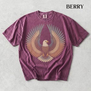 May include: A berry-colored short-sleeve t-shirt featuring a graphic of a bald eagle with its wings spread wide. The eagle is rendered in shades of brown, tan, and grey. The word BERRY is printed in black in the upper right corner of the image.