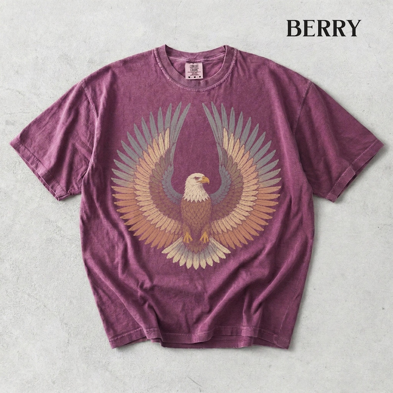 May include: A berry-colored short-sleeve t-shirt featuring a graphic of a bald eagle with its wings spread wide. The eagle is rendered in shades of brown, tan, and grey. The word BERRY is printed in black in the upper right corner of the image.