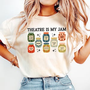 May include: Cream-colored t-shirt with the text "THEATRE IS MY JAM" above a graphic of nine jars. Each jar has a different label, including "Script Reading," "Props & Set," and "Acting Skills."