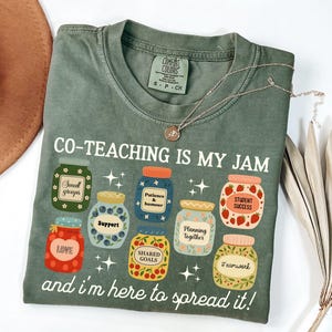 May include: A sage green t-shirt with the phrase "CO-TEACHING IS MY JAM" and various jar illustrations. The jars contain words like "Support" and "Teamwork". A silver necklace with a pendant is draped across the shirt.