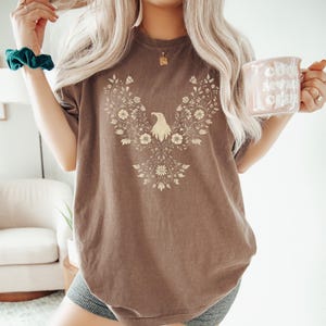 May include: Brown t-shirt featuring a floral eagle design. The eagle is centered on the shirt, surrounded by a detailed floral pattern. The shirt has a relaxed fit and is made of a soft material. The person is holding a mug that says "Cool Moms Club".