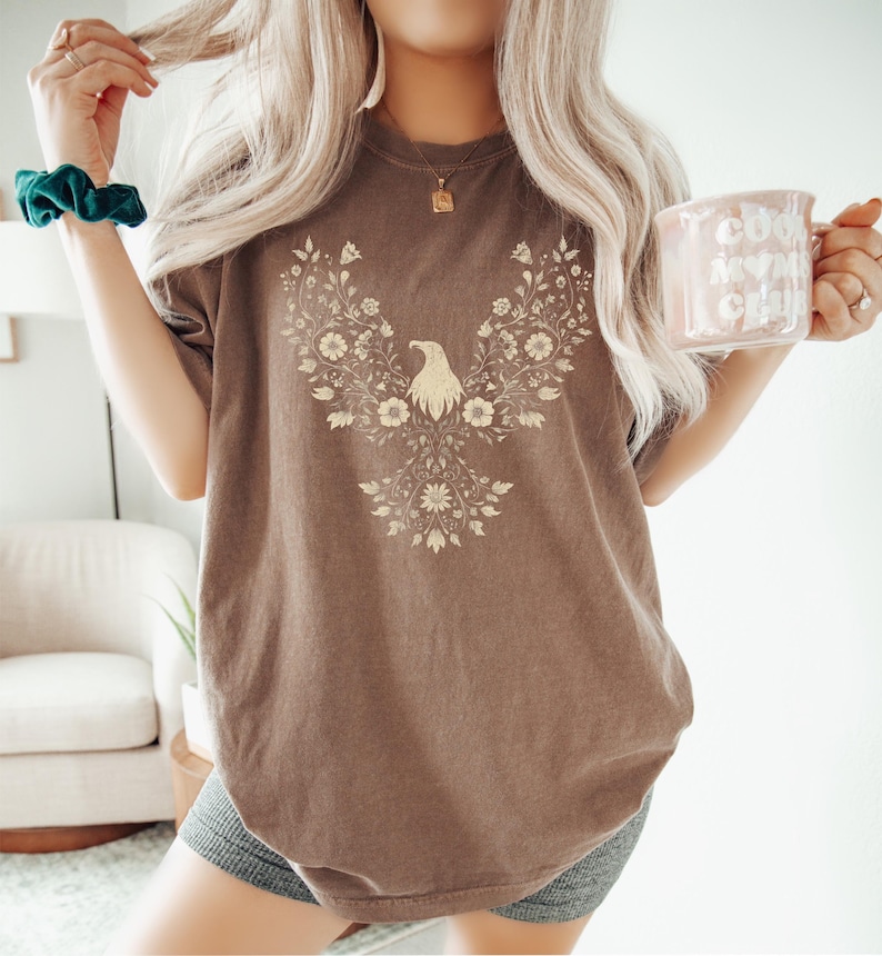May include: Brown t-shirt featuring a floral eagle design. The eagle is centered on the shirt, surrounded by a detailed floral pattern. The shirt has a relaxed fit and is made of a soft material. The person is holding a mug that says "Cool Moms Club".