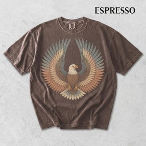 May include: A short-sleeve espresso brown t-shirt featuring a graphic of a bald eagle with wings spread wide. The eagle is rendered in muted tones of blue, tan, and brown. The word ESPRESSO is printed in black in the upper right corner of the image.