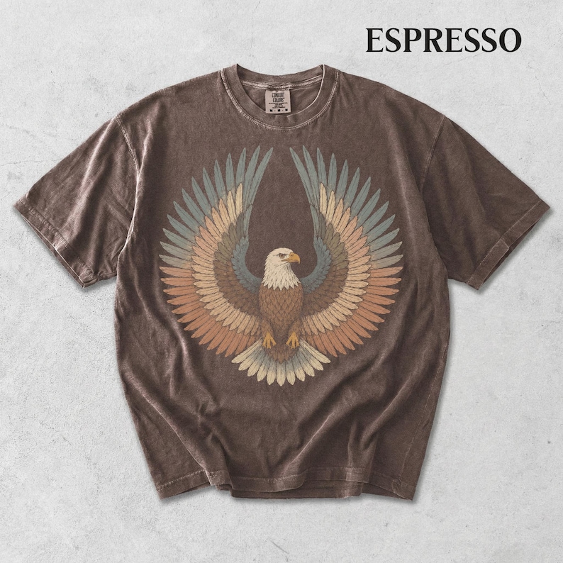 May include: A short-sleeve espresso brown t-shirt featuring a graphic of a bald eagle with wings spread wide. The eagle is rendered in muted tones of blue, tan, and brown. The word ESPRESSO is printed in black in the upper right corner of the image.