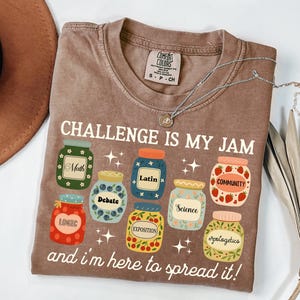 May include: Brown t-shirt with the text "CHALLENGE IS MY JAM" and various jar illustrations, each labeled with a different subject like Math, Debate, and Science. A silver necklace with a pendant rests on the shirt.