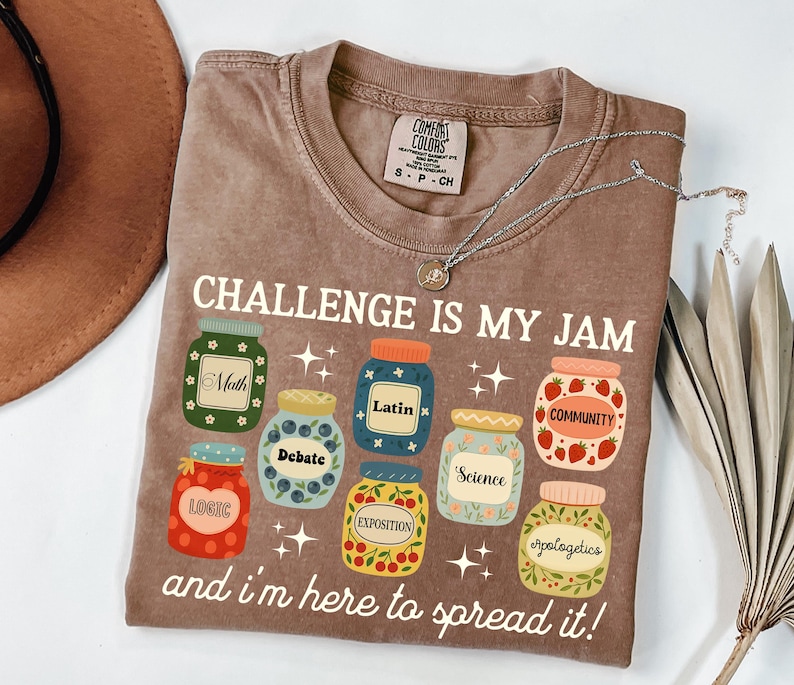 May include: Brown t-shirt with the text "CHALLENGE IS MY JAM" and various jar illustrations, each labeled with a different subject like Math, Debate, and Science. A silver necklace with a pendant rests on the shirt.