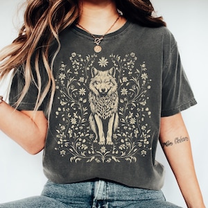 May include: Dark grey t-shirt featuring a detailed illustration of a wolf surrounded by floral elements. The wolf is in a centered, upright pose. The design is in a light beige color, creating a contrast against the dark grey fabric.