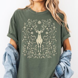 May include: Olive green t-shirt featuring a cream-colored deer design surrounded by floral elements. The deer has large antlers and stands centered within the floral frame. The shirt has short sleeves and a relaxed fit.