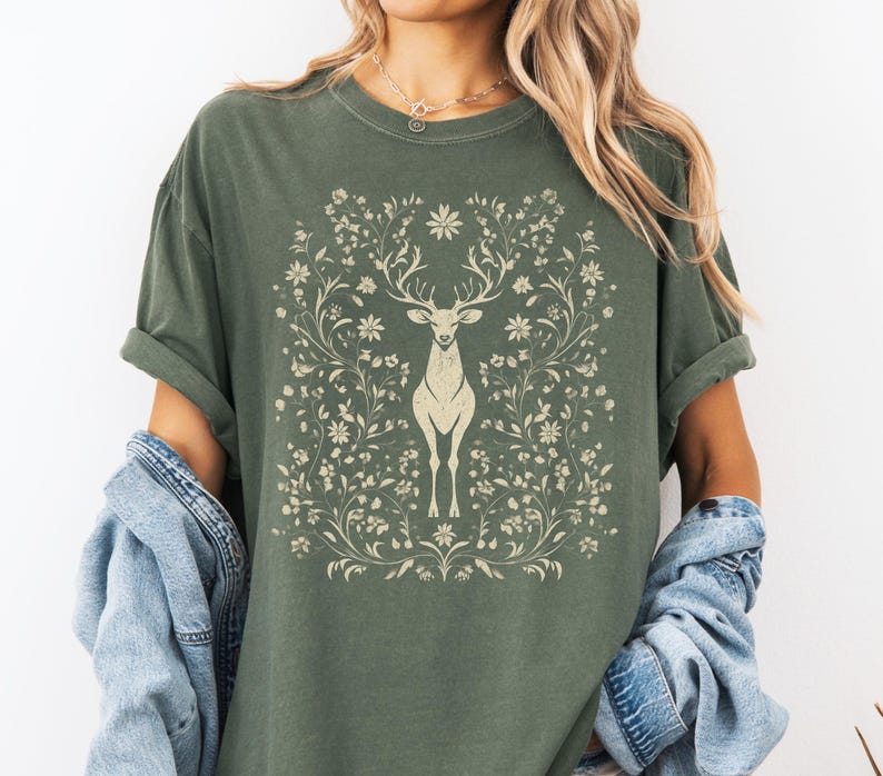 May include: Olive green t-shirt featuring a cream-colored deer design surrounded by floral elements. The deer has large antlers and stands centered within the floral frame. The shirt has short sleeves and a relaxed fit.