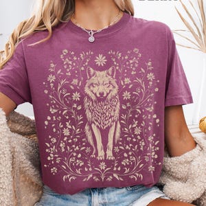 May include: A berry-colored short-sleeve t-shirt featuring a cream-colored graphic print of a wolf centered among intricate floral vines and blossoms. The text BERRY is printed in the upper right corner of the garment design.