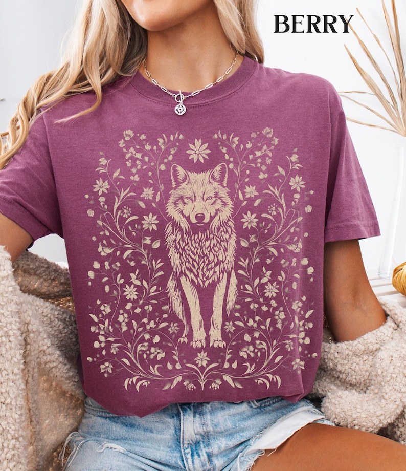 May include: A berry-colored short-sleeve t-shirt featuring a cream-colored graphic print of a wolf centered among intricate floral vines and blossoms. The text BERRY is printed in the upper right corner of the garment design.