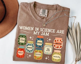 以下が含まれることがあります： 「WOMEN IN SCIENCE ARE MY JAM」の文字と、科学的なラベルが付いたジャムの瓶のイラストが描かれた茶色のTシャツ。シルバーのネックレスがTシャツの上にあります。Comfort Colorsブランド、サイズS-P-CH。