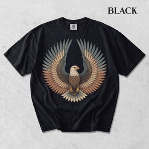 May include: A black short-sleeve t-shirt featuring a central graphic of a bald eagle with wings spread wide. The eagle's feathers are rendered in shades of brown, tan, and muted blue. The word BLACK is printed in the upper right corner of the image.