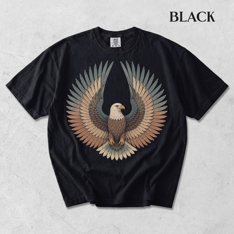 May include: A black short-sleeve t-shirt featuring a central graphic of a bald eagle with wings spread wide. The eagle's feathers are rendered in shades of brown, tan, and muted blue. The word BLACK is printed in the upper right corner of the image.