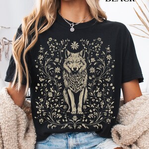 May include: A black short-sleeved t-shirt featuring a central graphic of a wolf surrounded by intricate floral vines and blossoms in a light beige color. The word BLACK is printed in the upper right corner of the image in a simple font.