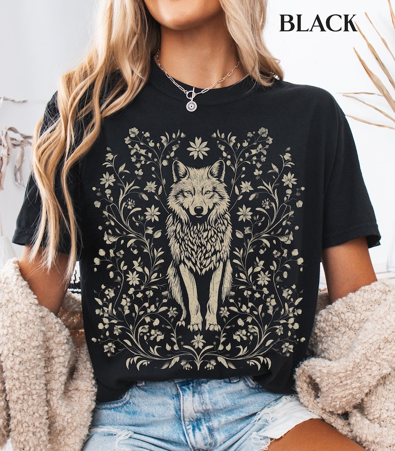 May include: A black short-sleeved t-shirt featuring a central graphic of a wolf surrounded by intricate floral vines and blossoms in a light beige color. The word BLACK is printed in the upper right corner of the image in a simple font.